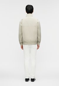 Ανοιχτό μπεζ μπουφάν με όρθιο γιακά, ελαστικά μανίκια και τελείωμα. Στη πλάτη διαθέτει οριζόντια ραφή και μια μικρή ετικέτα στο λαιμό.