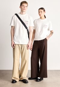 Valkoiset lyhythihaiset t-paidat, joissa painetut kuviot, paritettu beigeihin leveälahkeisiin housuihin ja ruskeisiin housuihin. Molemmilla henkilöillä mustat jalkineet.