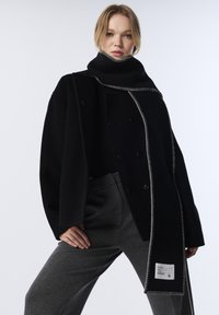 Abrigo negro de lana con cuello alto, que presenta costuras blancas en contraste y botones dobles, combinado con pantalones grises.