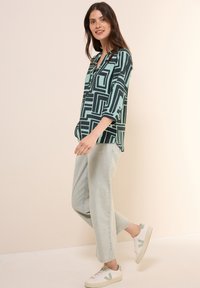 Groen en zwart gemusterde blouse met een V-hals en driekwart mouwen, gecombineerd met lichtdenim broek en witte sneakers met groene accenten.