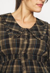 Chemise à carreaux marron et noirs avec un col à volants, une fermeture à boutons sur le devant et une taille froncée, ornée de lignes d'accent bleu dans le motif.