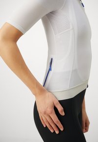 White mesh athletic top with short sleeves, featuring blue zipper details and a fitted design. Paired with black leggings. Textured fabric.