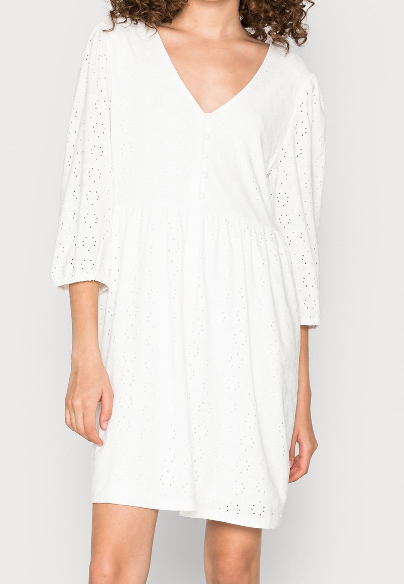 Robe blanche avec un décolleté en V, des manches bouffantes et une taille froncée. Présente des détails en œillets et des accents de boutons sur le devant.