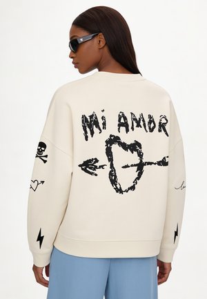 Donna con occhiali da sole indossa una felpa color panna con una grafica nera di "Mi Amor," cuore e freccia, teschio e fulmine sul retro e sulle maniche.