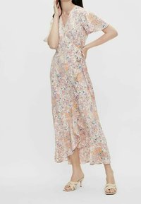 Femme portant une robe midi à fleurs avec des manches volantées et des sandales à talons beiges, posant avec une main sur la hanche et l'autre touchant son visage.