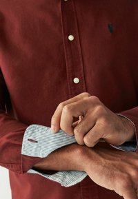 Chemise rouge à boutons avec un poignet rayé bleu et blanc à motif. Présente un petit logo sur la poitrine gauche et des boutons sur le devant.