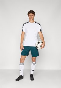 Camiseta deportiva blanca con rayas negras, pantalones cortos azul verdoso, botas negras y calcetines blancos con rayas negras. Sosteniendo un balón de fútbol estampado.