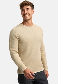 Pull en tricot beige avec un motif texturé, col rond et manches longues. L'ourlet présente un détail de finition blanc contrastant.