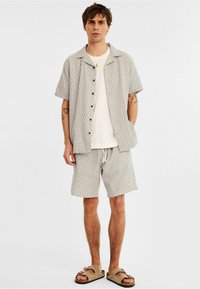Chemise à manches courtes à boutons et shorts assortis en tissu gris clair avec un motif en losanges, associés à des sandales à glissière beiges.