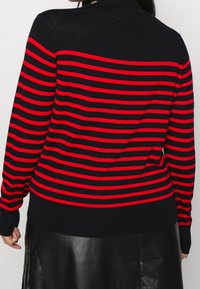 Maglione a righe nere e rosse con colletto alto, maniche lunghe e polsini a coste. Il design presenta ampie righe orizzontali sulla parte posteriore.