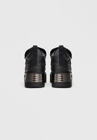 Tenis pretos com um cabedal texturizado, apresentando uma entressola grossa e brilhante na cor preta, calcanhar reforçado e ilhós metálicos para os atacadores.
