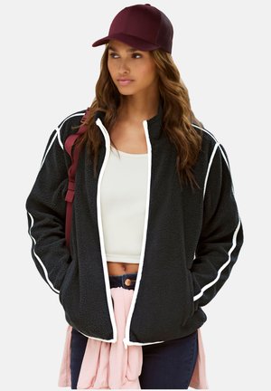 Junge Frau, die eine schwarze Fleecejacke mit weißem Rand, ein weißes Crop-Top, dunkle Jeans, eine kastanienbraune Mütze und einen pinkfarbenen Pullover um die Taille gebunden trägt.