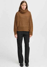 Pull en tricot marron avec un col montant, associé à un pantalon noir droit. Présente des poignets côtelés et une coupe décontractée.