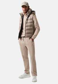 Beige puffer vest met een capuchon over een bijpassende hoodie en broek. Witte sneakers met grijze accenten. Bevat ritszakken en een trekkoord.