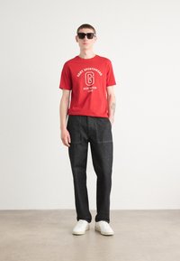 Rød t-shirt med hvid branding og tekst; sorte denimbukser med synlige sømme; hvide sneakers; solbriller; neutral baggrund.