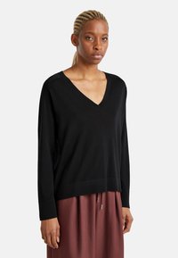 Maglione nero a scollo a v realizzato in un materiale morbido, con maniche lunghe e una vestibilità rilassata; presenta un orlo a coste e spacchi laterali, indossato su una gonna marrone.