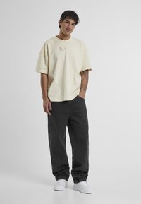 Ülikõrge beež puuvillane T-särk kuldse logoga, millele on lisatud mustad teksapüksid ja valged tossud. Modell seisab lihtsa tausta ees.