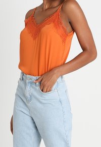 Orange satin cami-top med spetsdetaljer vid halsringningen. Modellen bär ljusblå högmidjade jeans som framhäver toppens strukturerade tyg.