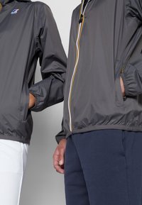 Des vestes grises légères avec des poignets élastiques, des fermetures éclair avant avec des accents colorés et des poches latérales, associées à des pantalons bleu marine et blancs.