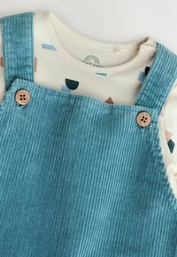 Blaue Cord-Dungarees mit Holzknöpfen, darüber ein cremefarbenes Hemd mit einem geometrischen Muster in Pink, Grün und Blau.