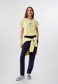 Femme debout portant un t-shirt jaune, un pantalon de jogging bleu marine, un sweat-shirt jaune noué à la taille, des baskets argentées et des boucles d'oreilles créoles sur un fond uni.