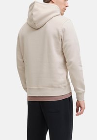 Beige hætte sweatshirt med en blød tekstur, synlig syning og en korteret design. Båret over et pink lag og parret med sorte bukser.