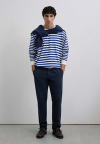 Cămașă cu dungi pe mânecă lungă în albastru și alb, cu un pulover bleumarin aruncat pe umeri, asortată cu pantaloni bleumarin și pantofi închiși la culoare.