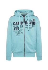 Light blue zip-up hoodie made of soft fabric. Features a hood, front pockets, and printed graphics in dark blue and black. Plain cuffs and hem.