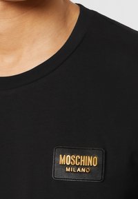 Svart t-shirt med rund halsringning, med en rektangulär svart läderlapp med guldtext som säger "MOSCHINO MILANO."