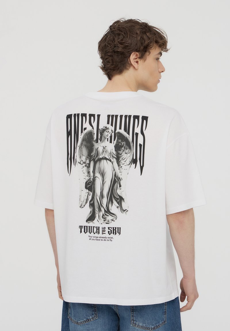 T-shirt bianco oversize con una grande grafica di un angelo nero sul retro, con il testo "ALI D'ANGOLO" e "TOCCA IL CIELO" sottostante.