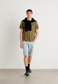 Olivengrønn t-skjorte, svart genser drapert rundt halsen, lyseblå denimshorts, svarte sokker, svarte dressko, nøytral bakgrunn.