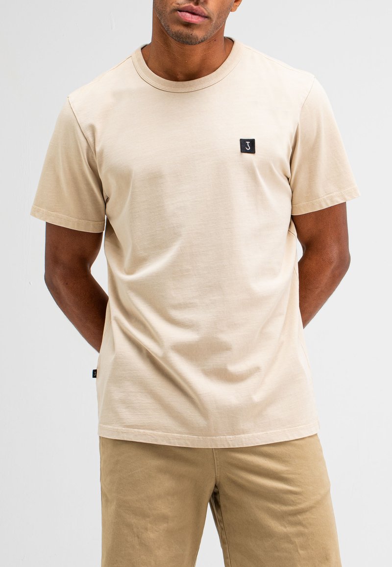 Lichtbeige T-shirt van zachte stof, met een ronde hals, korte mouwen en een klein zwart logopatch aan de voorkant.