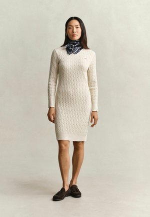Frau trägt ein cremefarbenes, kabelgestricktes Langarmkleid, einen dunkelblau gestreiften Halsschal und dunkelbraune Loafers, stehend vor einem schlichten Hintergrund.