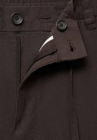 Braune Hose mit Reißverschluss, ausgestattet mit schwarzen Knöpfen und einem strukturierten Stoff. Sichtbares weißes Gewebe im Bereich des Reißverschlusses.