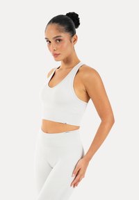 Γυναίκα με κότσο φορώντας λευκό αθλητικό crop top και ψηλόμεσο κολάν που ταιριάζουν, στέκεται μπροστά σε απλό φωτεινό φόντο.