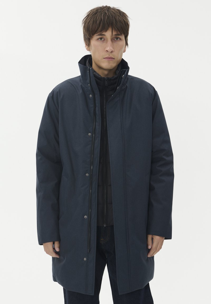 Jauns vīrietis, tērpies tumši zilā pildītā ziemas mētelī, virsū melnā jaciņā ar rāvējslēdzēju, stāv pretī vienkāršai gaišai fona.