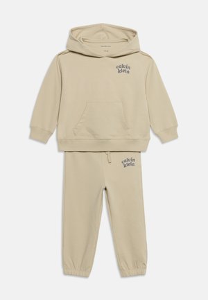 Beżowy zestaw dziecięcej bluzy z kapturem i spodni dresowych Calvin Klein z kieszeniami z przodu i nadrukiem logo na piersi oraz górnej części uda.