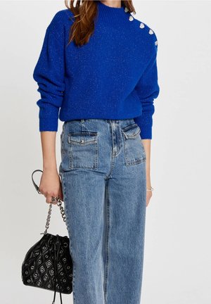 Femme portant un pull bleu vif avec des boutons sur les épaules, un jean bleu taille haute avec des poches avant, tenant un sac à main noir matelassé.