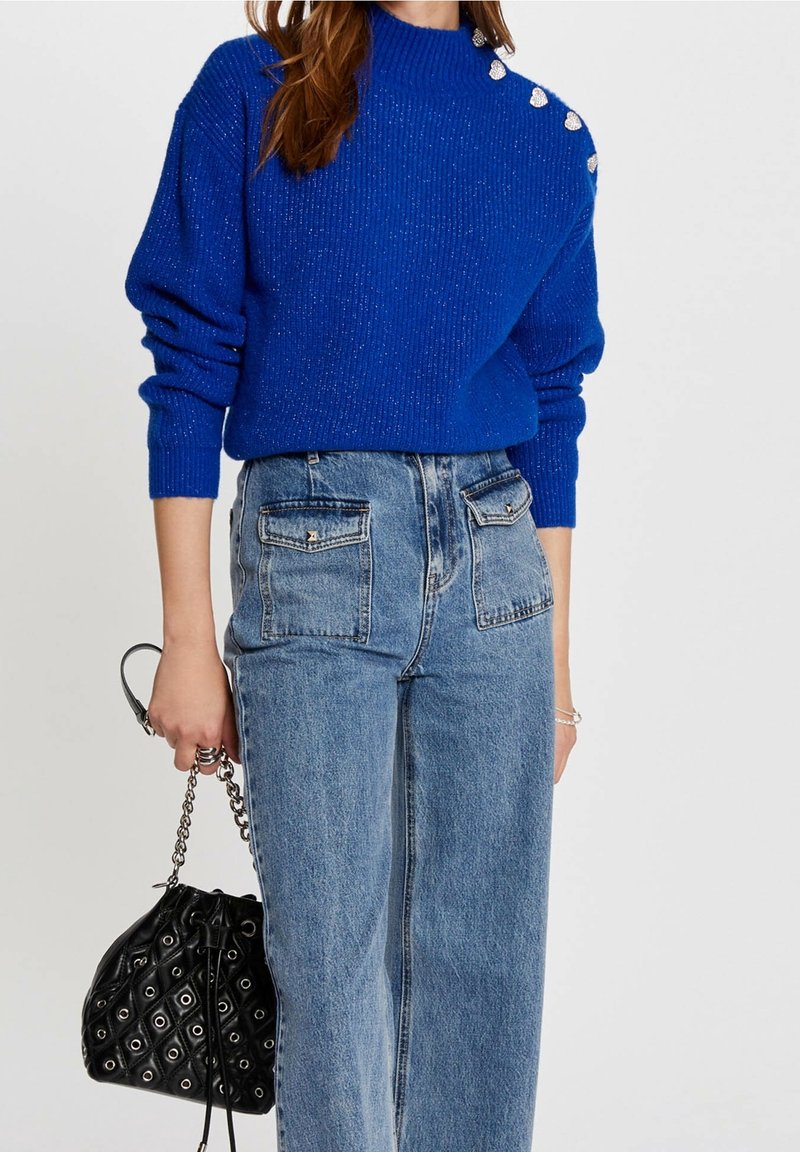 Femme portant un pull bleu vif avec des boutons sur les épaules, un jean bleu taille haute avec des poches avant, tenant un sac à main noir matelassé.