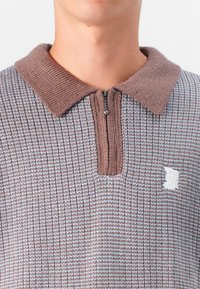 Knit polo shirt in brown and light blue, features a zip-up collar and a textured ribbed pattern with a small white logo patch on the chest.