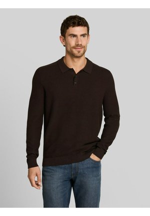 Mann mit kurzen lockigen Haaren trägt einen schwarzen Polo-Pullover mit langen Ärmeln und blaue Jeans, steht mit einer Hand in der Tasche vor einem grauen Hintergrund.