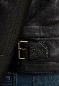 Črna usnjena jakna z teksturirano površino, ki ima kovinski sponko, pod kotom pripet trak in kontrastno šivanje na robu.