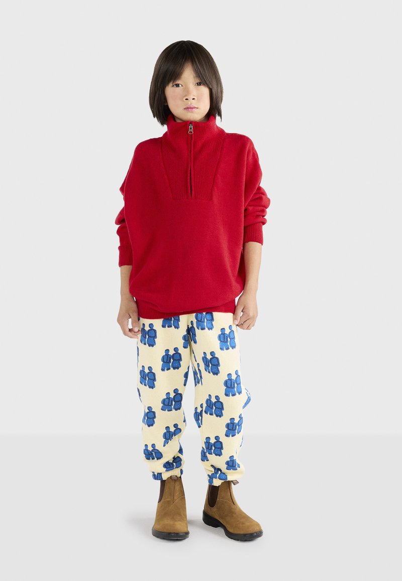 Rød strikket fleece-pullover med høj krave, parret med cremede bukser med blå menneskefigur-mønstre og brune ankelstøvler.