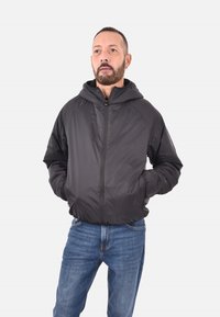 Giacca nera leggera, con zip e cappuccio. Presenta polsini elasticizzati e tasche laterali. Indossata sopra jeans blu. Materiale liscio e resistente all'acqua.