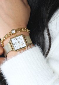 Gold mesh watch with a square white face and gold Roman numerals, paired with a gold chain bracelet featuring a round stone accent.