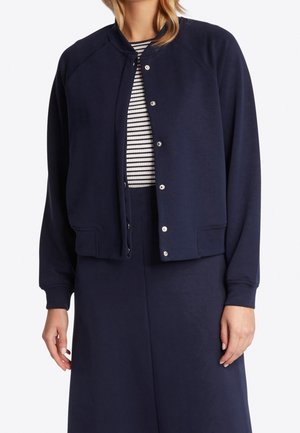 Femme portant une veste bleu marine à boutons sur un haut à rayures noires et blanches, assortie à une jupe ou un pantalon bleu marine, debout devant un fond blanc.