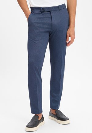 Mann steht mit den Händen in den Taschen, trägt schlank geschnittene blaue Anzughosen und schwarze Slip-On-Schuhe mit weißen Sohlen vor weißem Hintergrund.