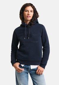 Felpa blu navy con cappuccio e coulisse, caratterizzata da un logo GANT in rilievo e dalla scritta "American Sportswear". Tessuto misto cotone con vestibilità comoda.