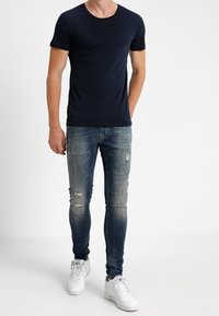 Man som bär en åtsittande svart t-shirt, slitna smala blå jeans och vita sneakers, stående mot en enkel vit bakgrund.