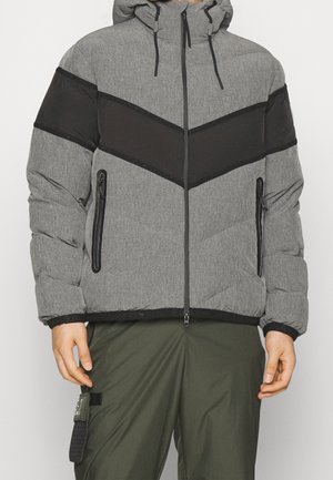 Homme portant une veste matelassée grise avec une bande chevron noire, fermeture éclair frontale, capuche et poches latérales zippées, assortie à un pantalon vert olive.